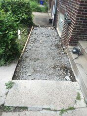 Gravel walkway under construction next to a brick building. Concrete slab at the bottom, grass and bushes on the left.