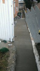 Newly poured concrete sidewalk next to a white picket fence, leading toward a building.