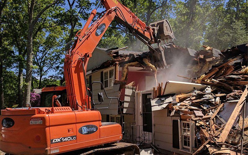 Orange excavator demolishing a house amid debris and dust under trees