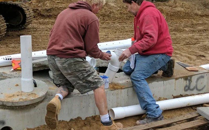 Two workers in hoodies and gloves install white pipes on a concrete utility base outdoors.