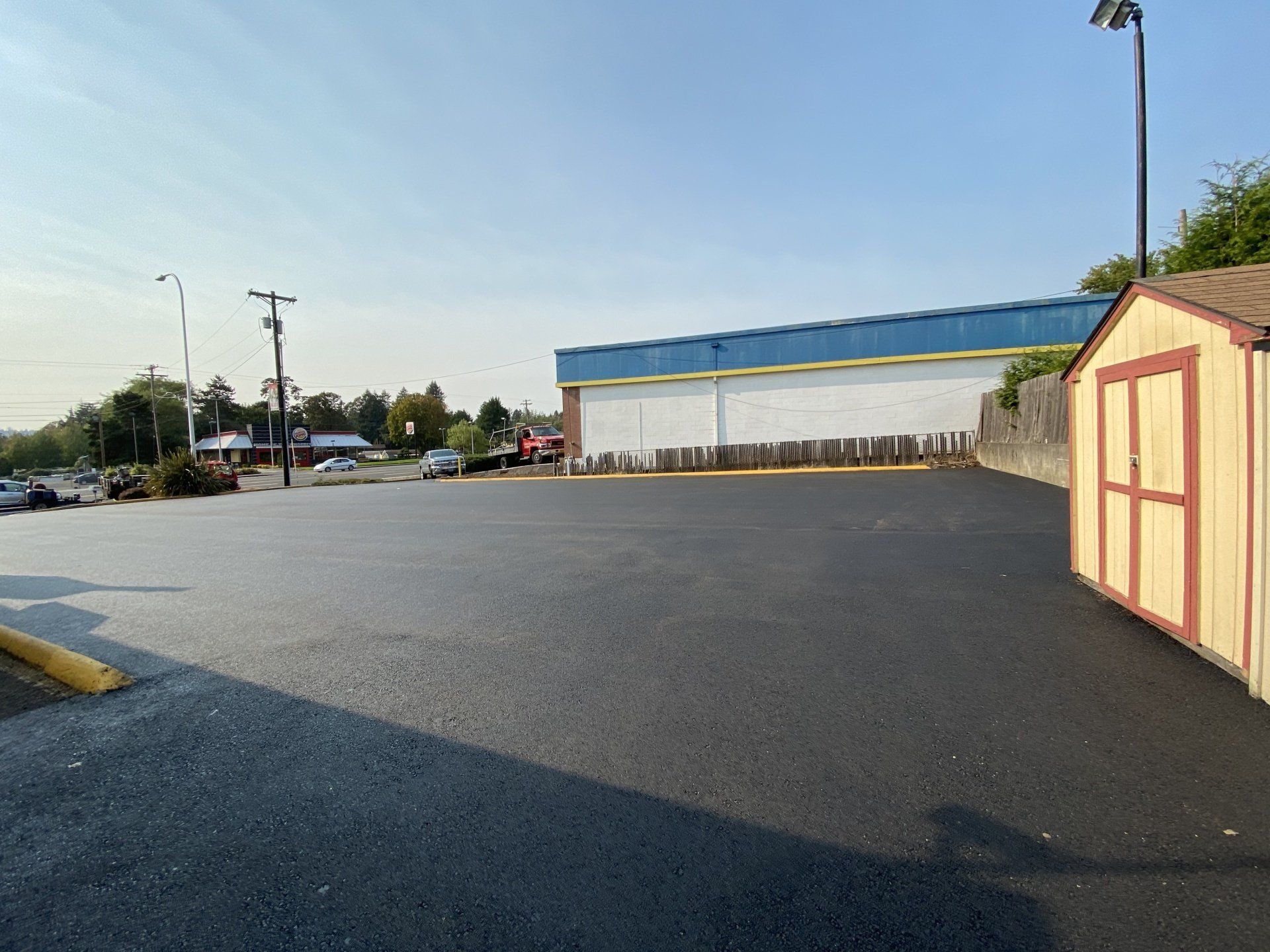 Empty asphalt parking lot with a building and shed in the background under a blue sky.