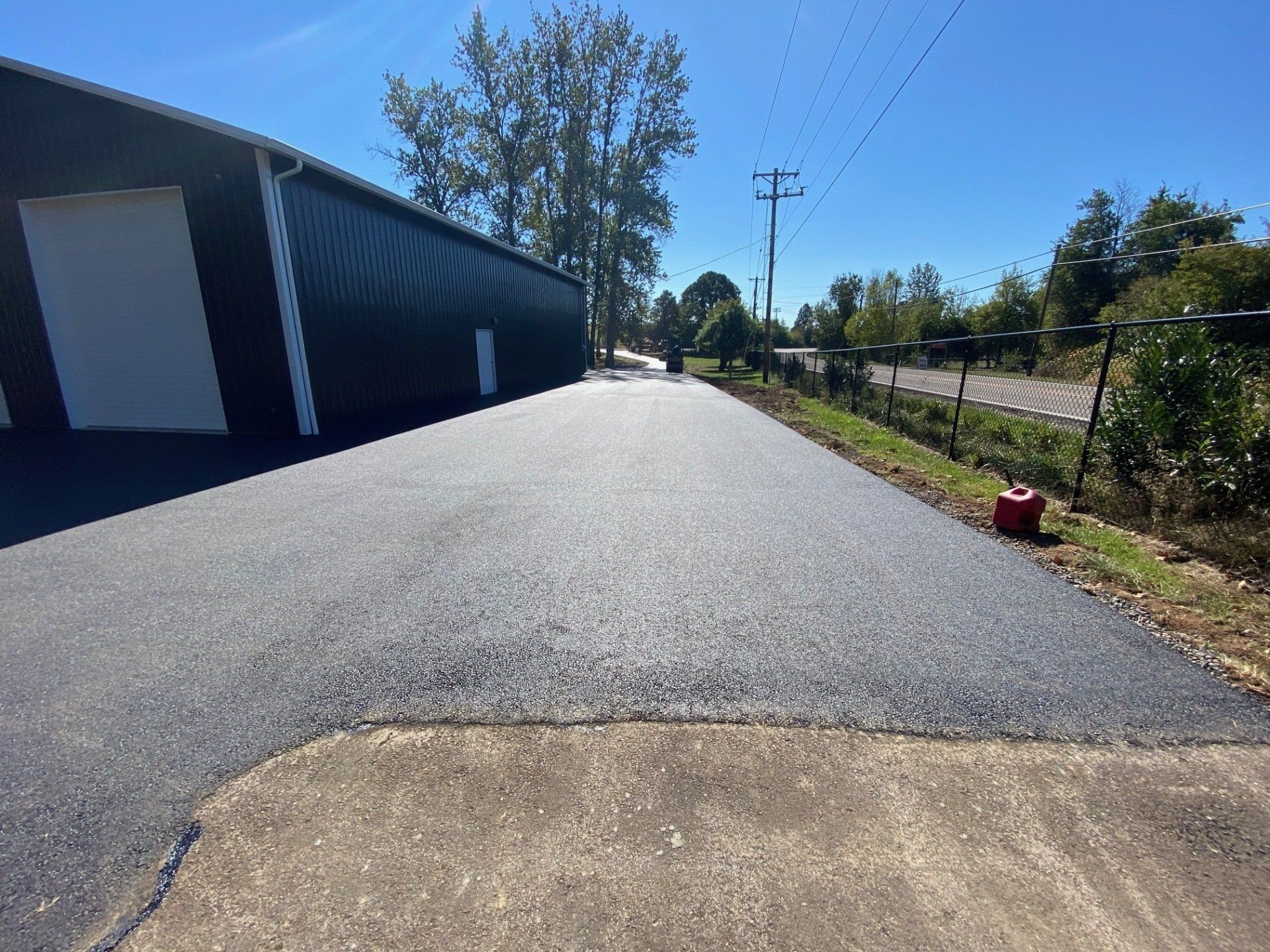 Asphalt driveway next to a black building under a blue sky, with utility poles and a fence in the distance.