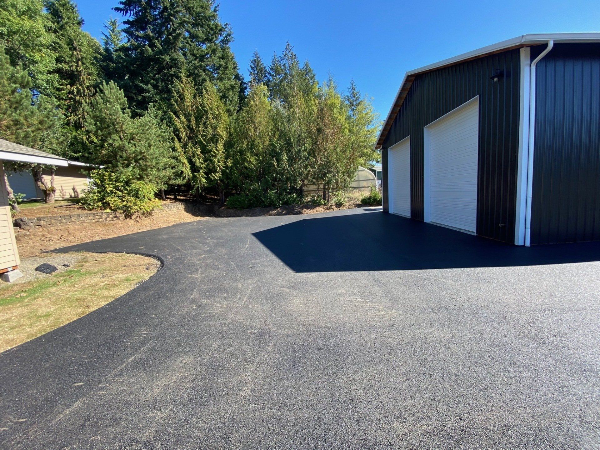Asphalt driveway curves to a dark metal building with white garage doors, trees, and blue sky.