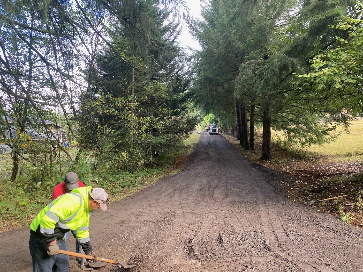 Workers spreading gravel on a road lined with trees. One wears a reflective vest, the other a hat.