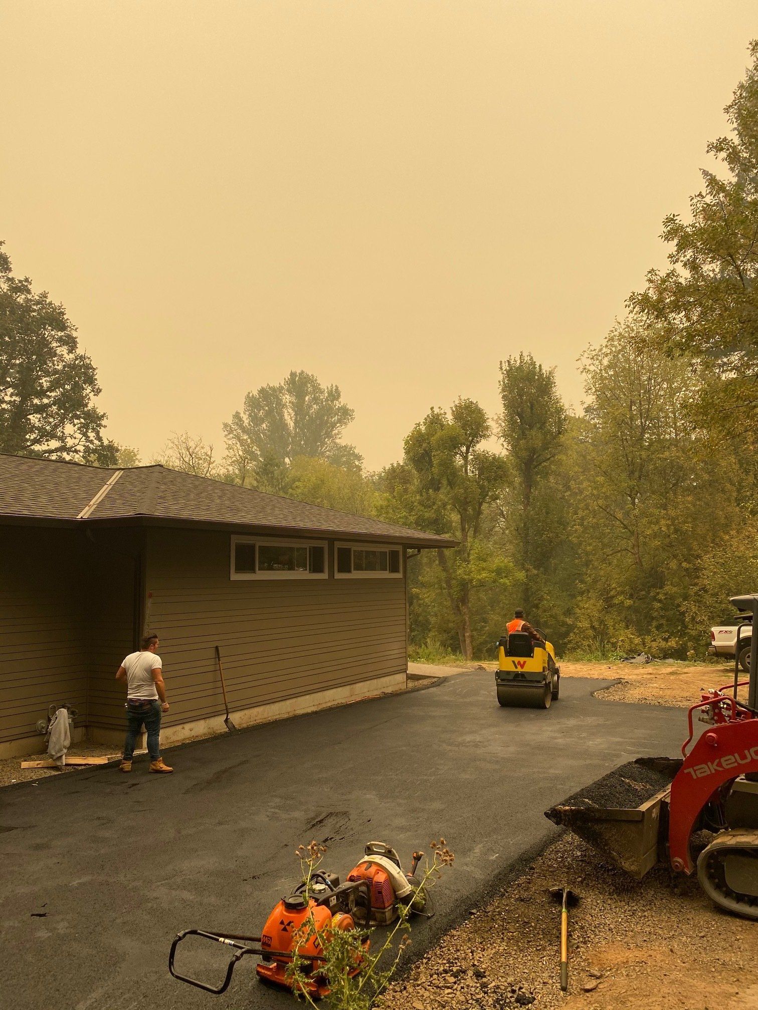 Asphalt paving in progress. Worker near building, roller compacting, sky hazy with smoke.