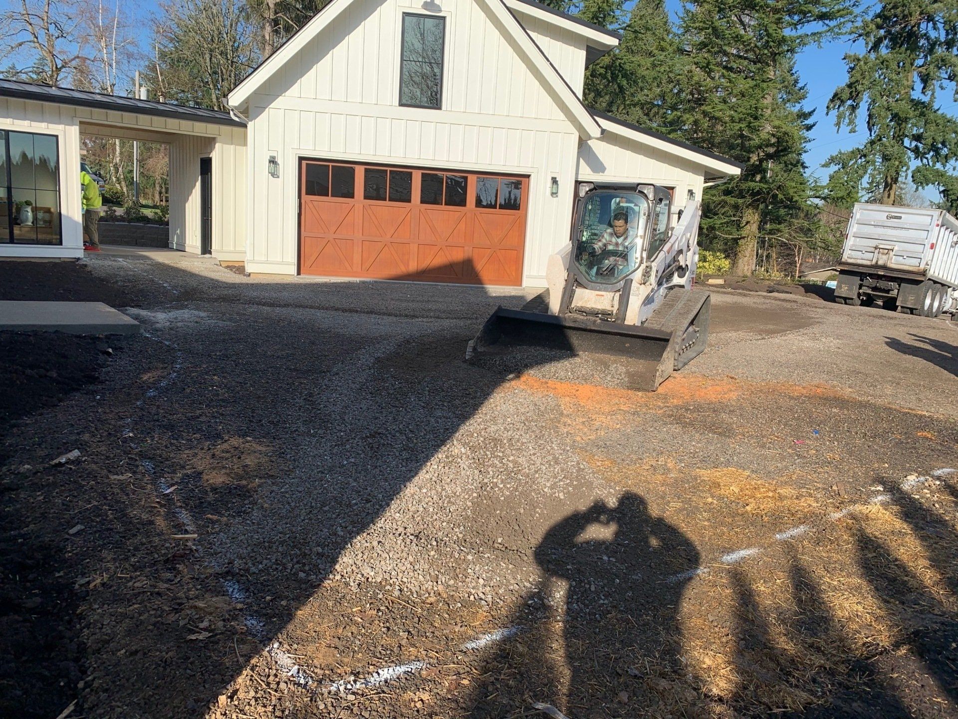 A small white building with a brown garage door under construction, Bobcat, and gravel driveway.