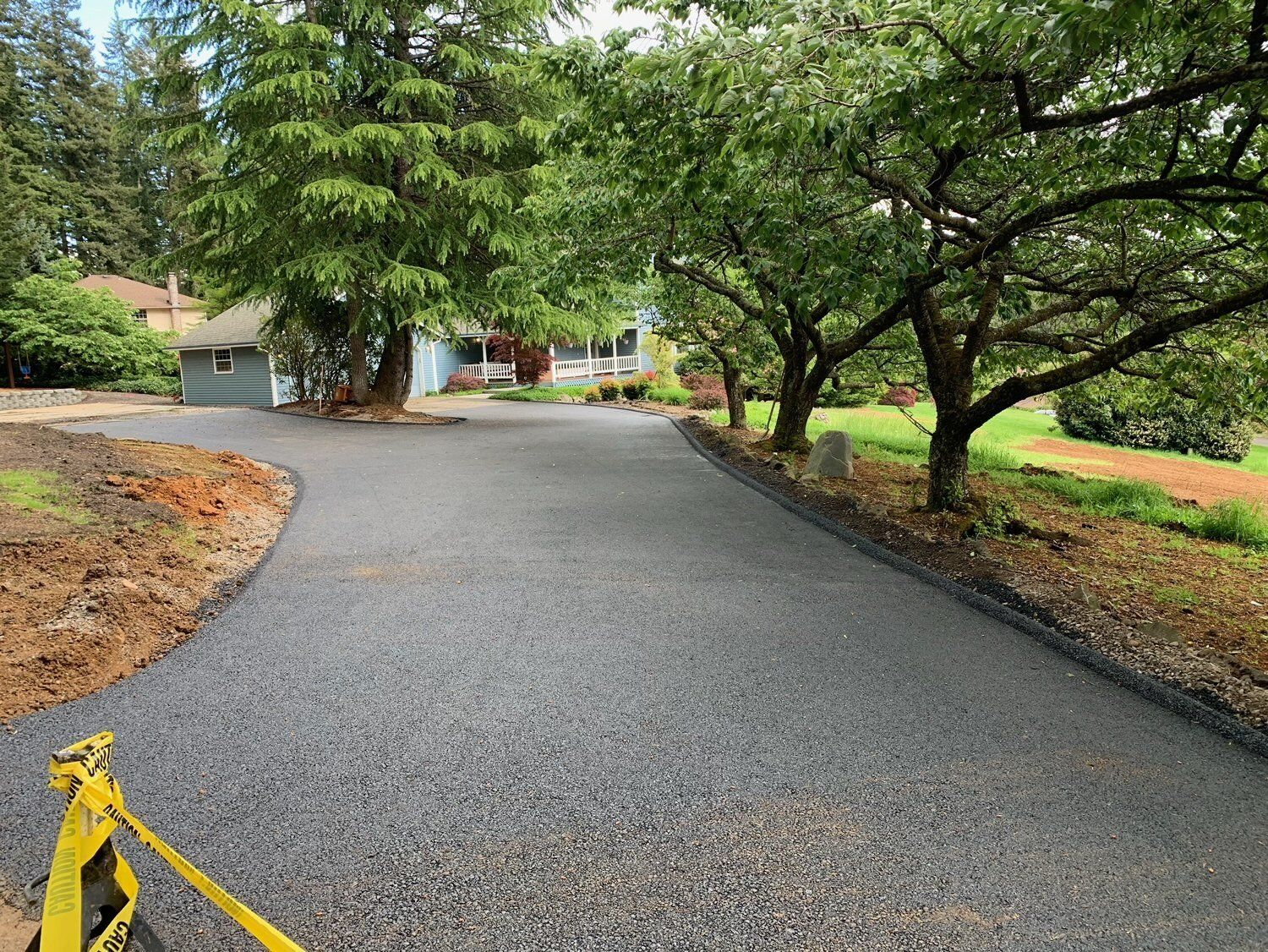 Asphalt driveway with trees lining the right side, leading to a house. Yellow caution tape in the foreground.