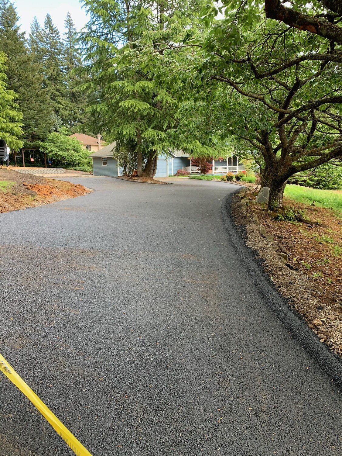 Gravel driveway curves toward a blue house, under large trees. Yellow caution tape in the lower left corner.