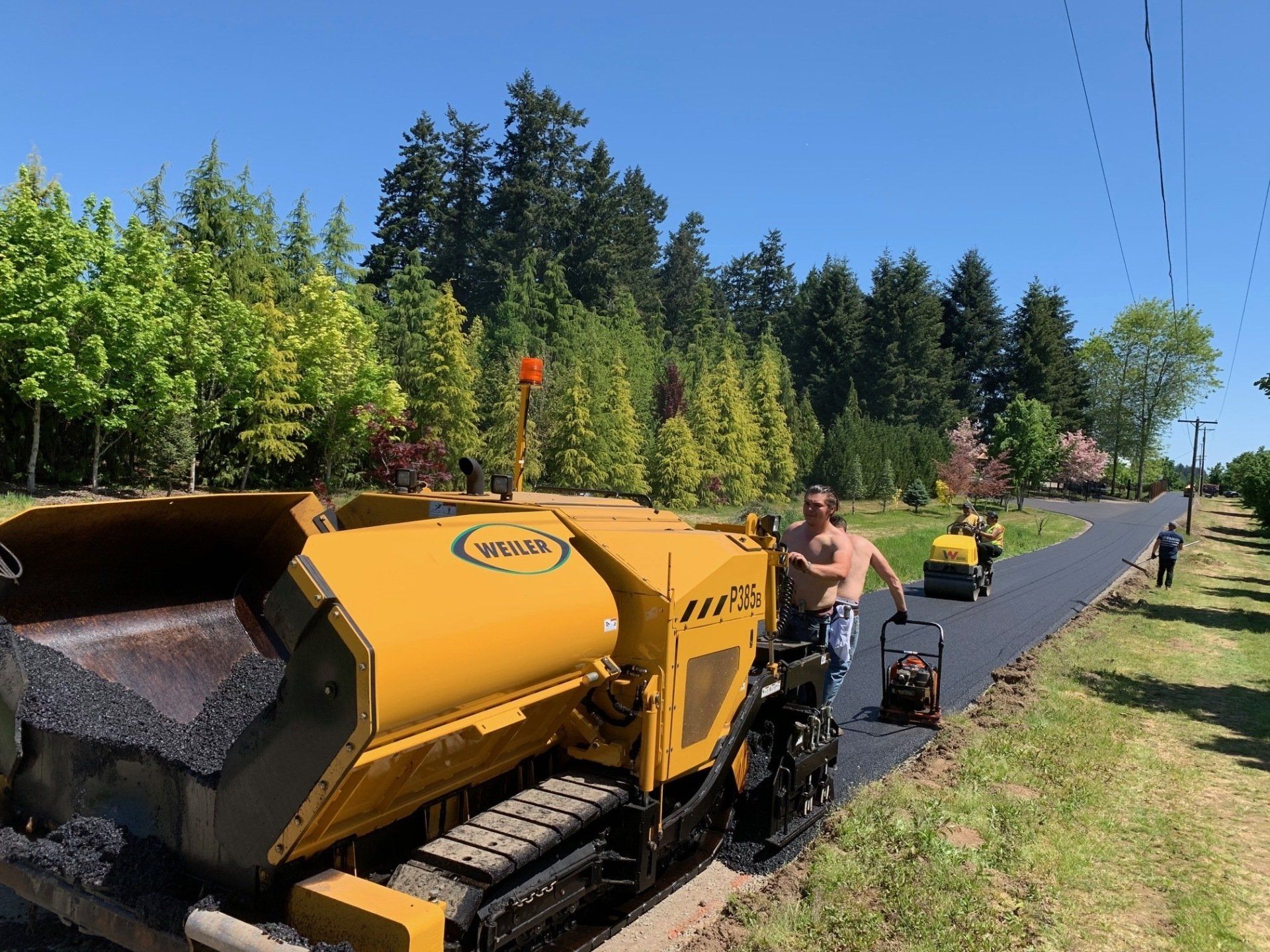 Road paving crew laying asphalt; yellow machine, man operating roller, trees in background.