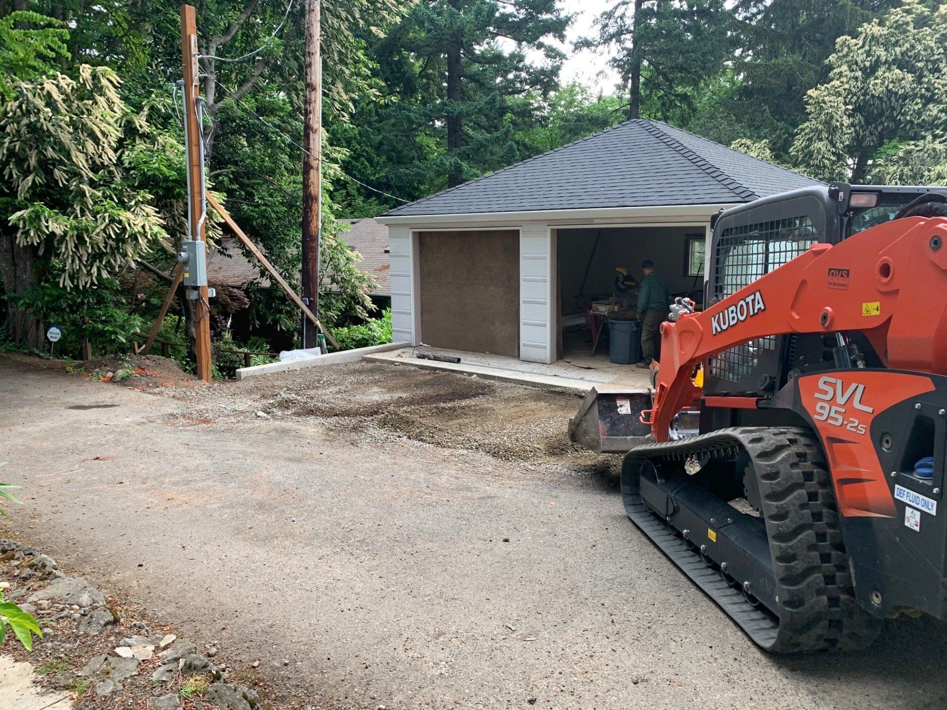 Orange Kubota skid steer grading gravel driveway in front of a two-car garage.