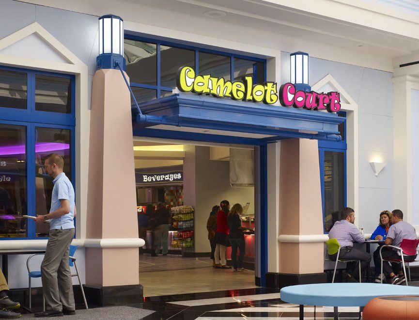 Entrance to Camelot Court restaurant with blue and pink accents. People are dining at tables outside; a man stands in the doorway holding papers.