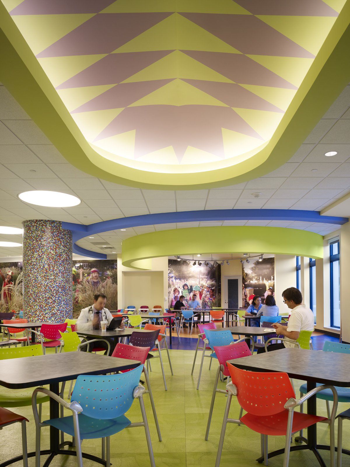 Colorful cafeteria with round tables and chairs. Pastel ceiling with geometric pattern. People sit and eat.