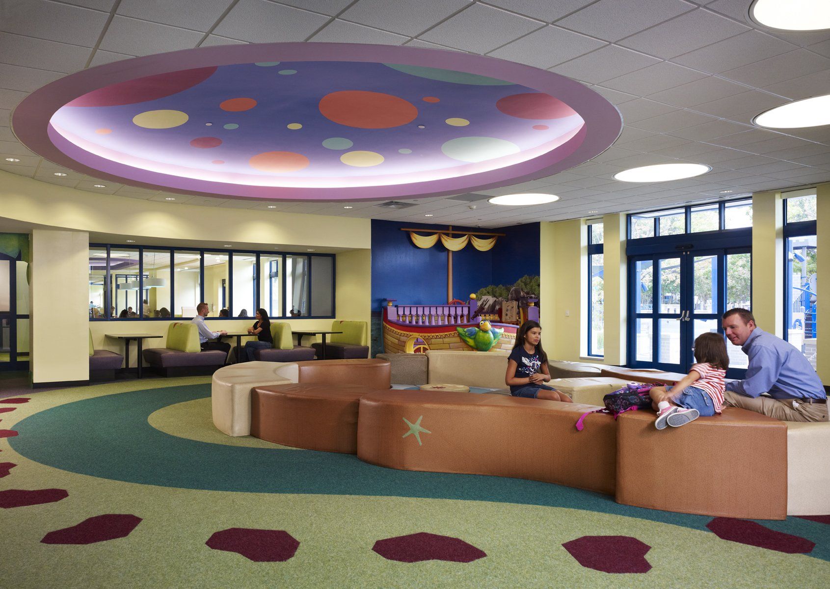 A colorful children's library area with a circular seating area, a boat-shaped play structure, and parents with children.