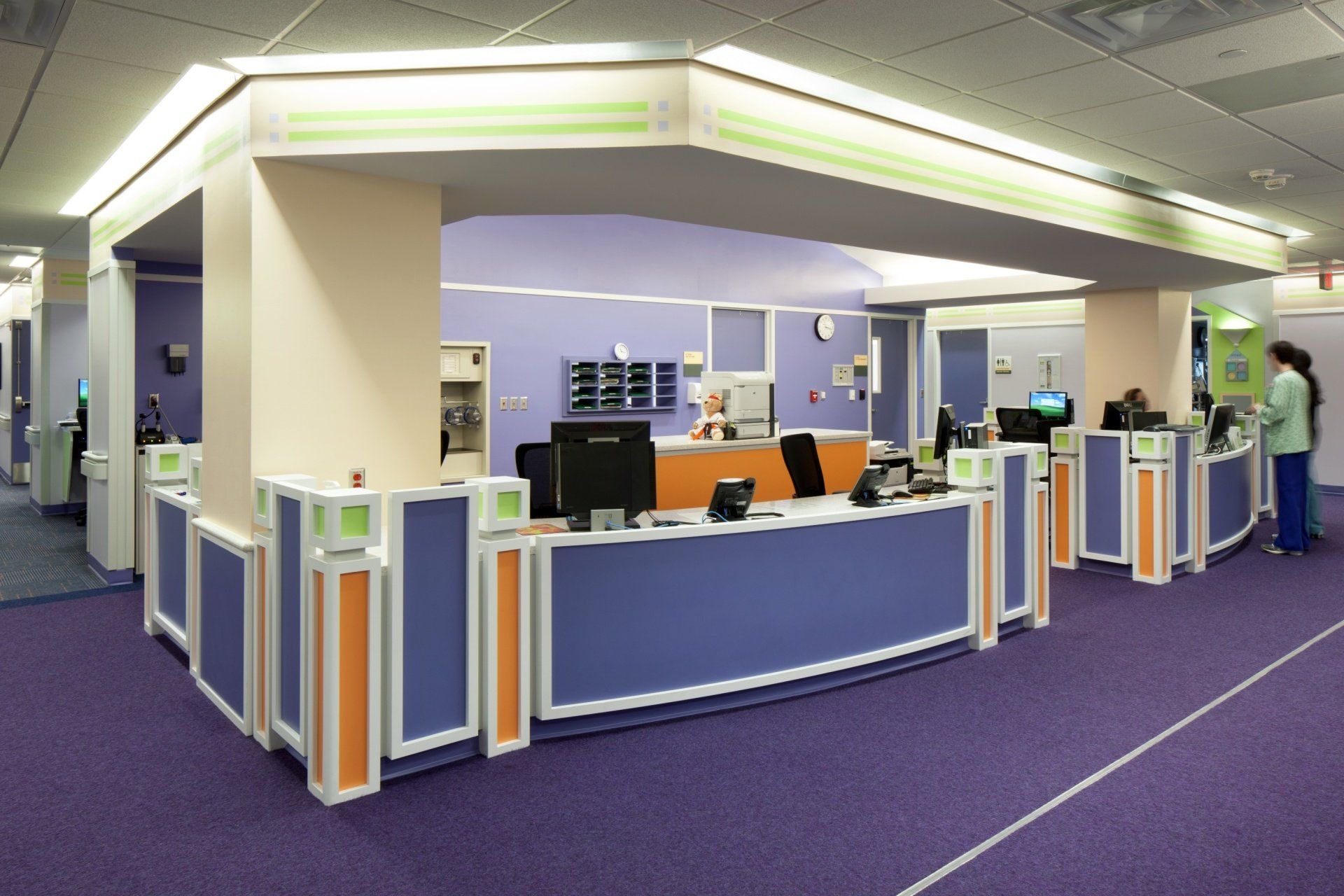 A hospital reception desk with purple carpet, blue and orange accents, and a worker in scrubs.