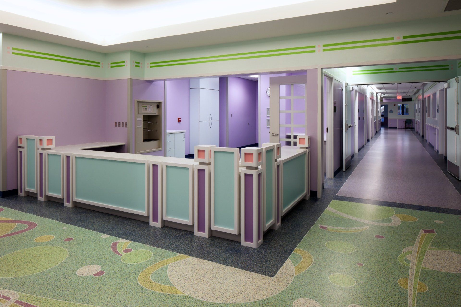 Reception desk in a hospital hallway with pastel colors, including lavender walls and a mint-colored desk. A long hallway extends to the right.