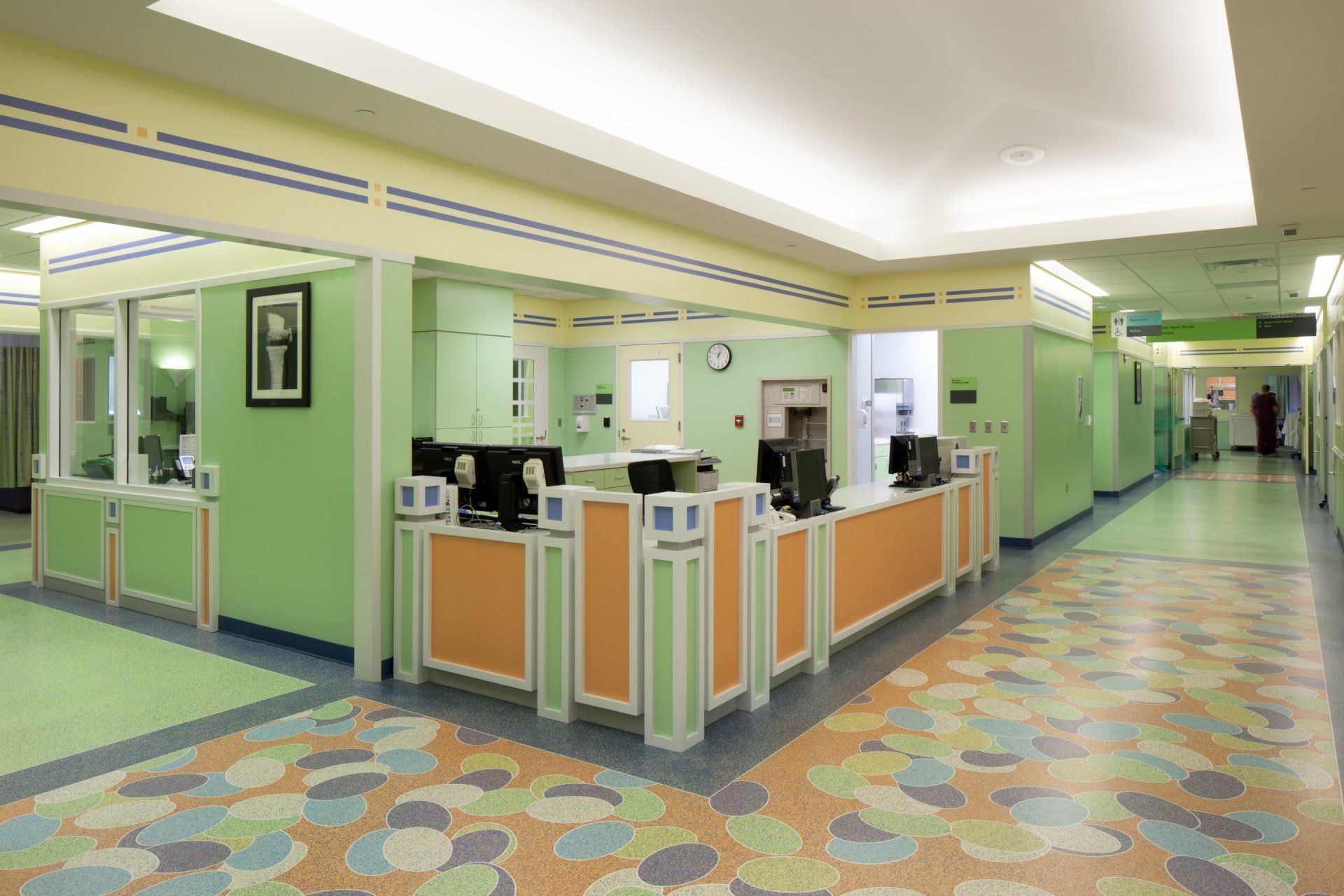 Hospital reception desk and hallway with pastel green and orange accents, patterned flooring, and a person walking down the hallway.