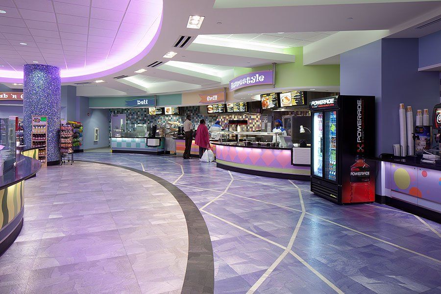 Interior of a movie theater lobby with concession stands and curved flooring. Purple and blue dominate the color scheme. People are visible at the counters.