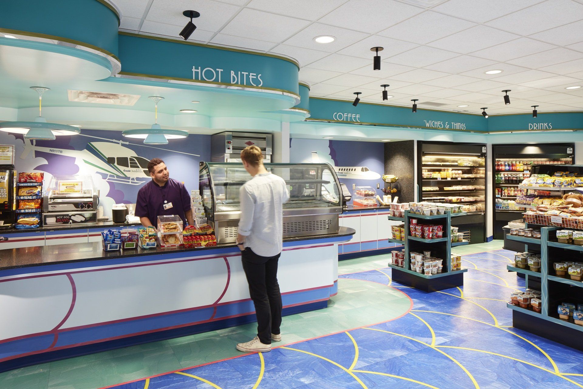 A man serves a customer at a brightly-colored cafe counter. Blue and white decor, with snacks and drinks on display.