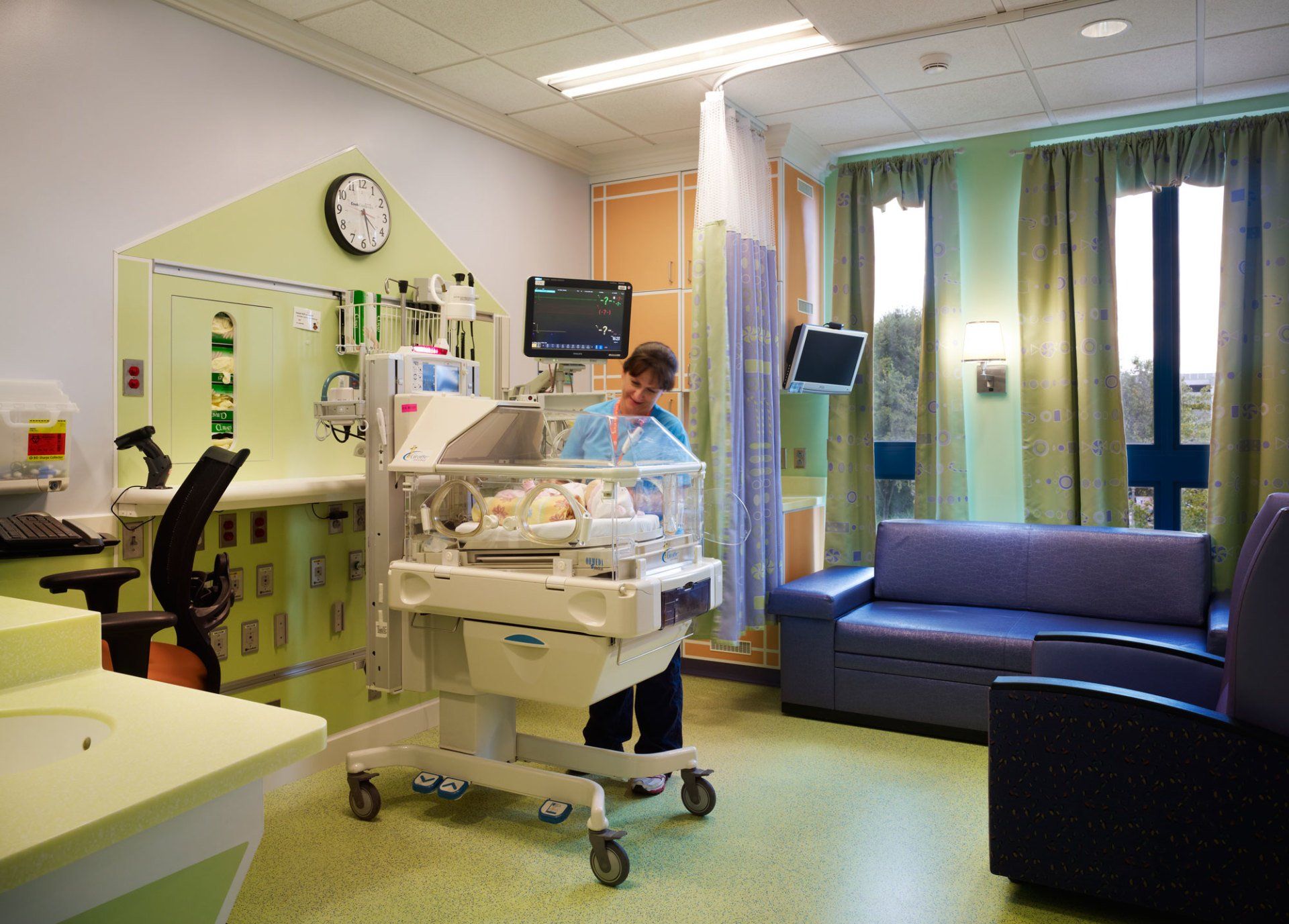 A medical professional tending to a newborn in an incubator within a hospital room; a clock and equipment are mounted on the wall.