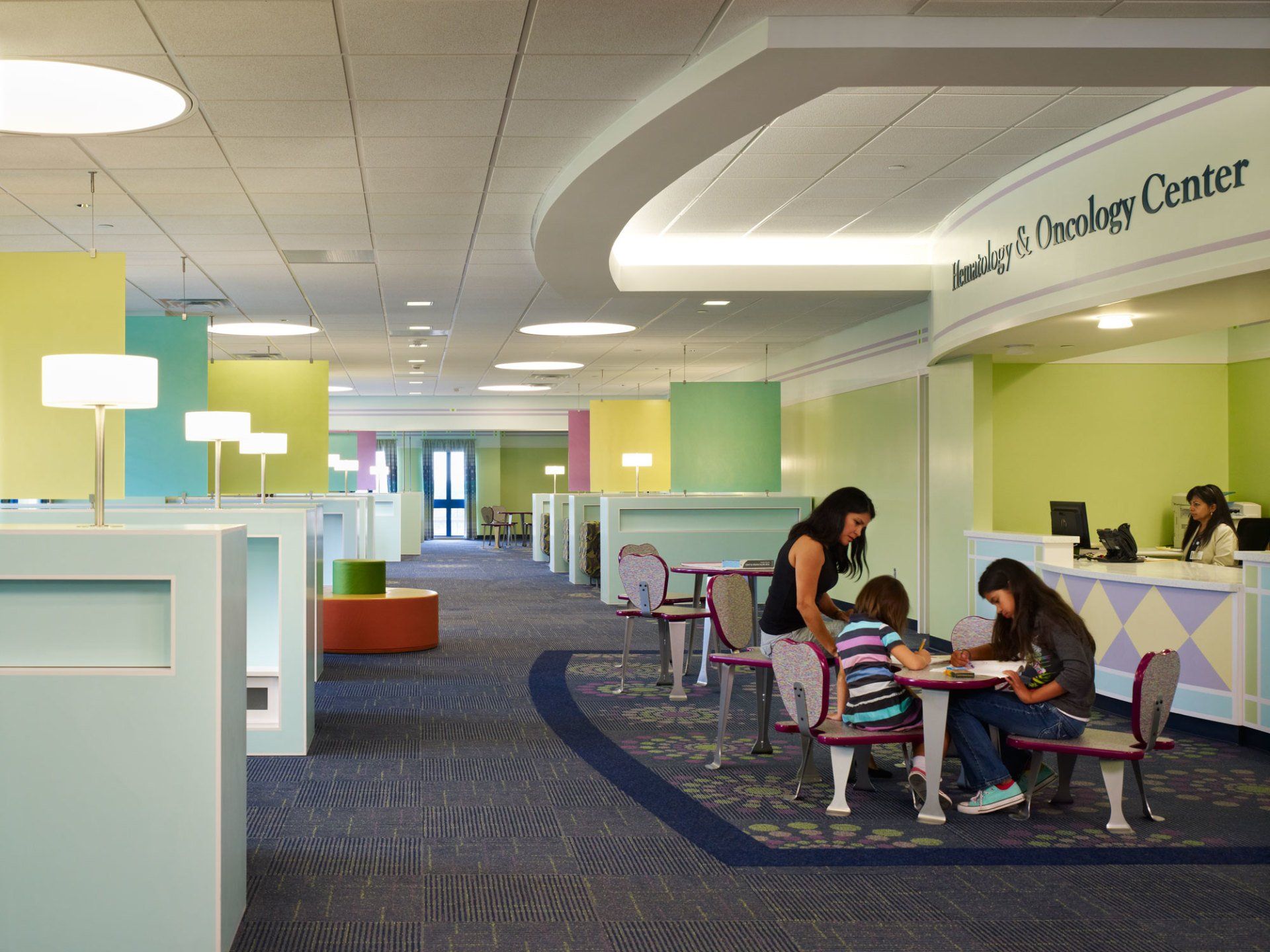Interior of a brightly colored medical center waiting area. Two women and a child are seated at a table, and two staff members are at a counter.
