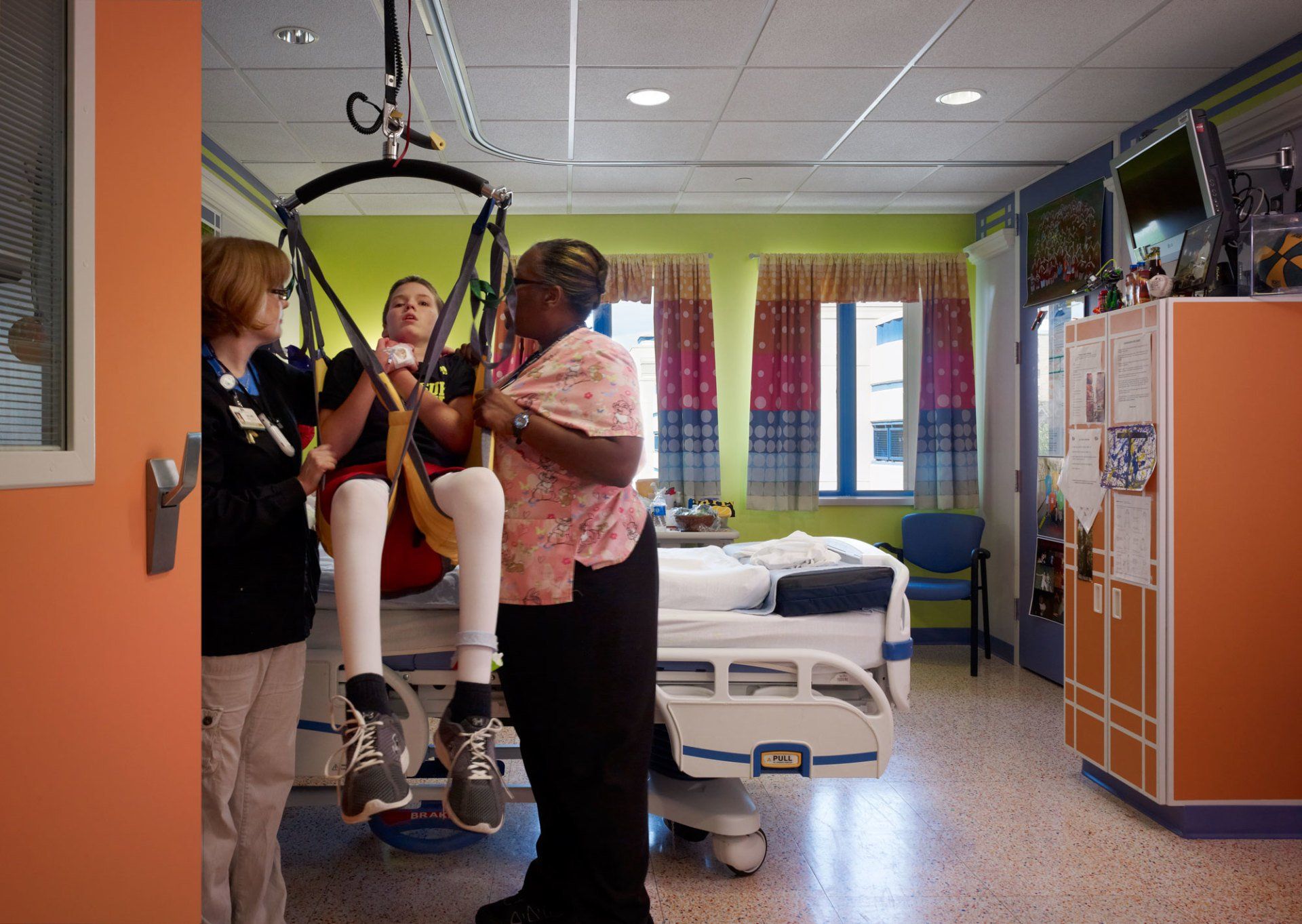 Three caregivers assisting a young person suspended in a ceiling lift in a hospital room. The room has colorful decor and a hospital bed.