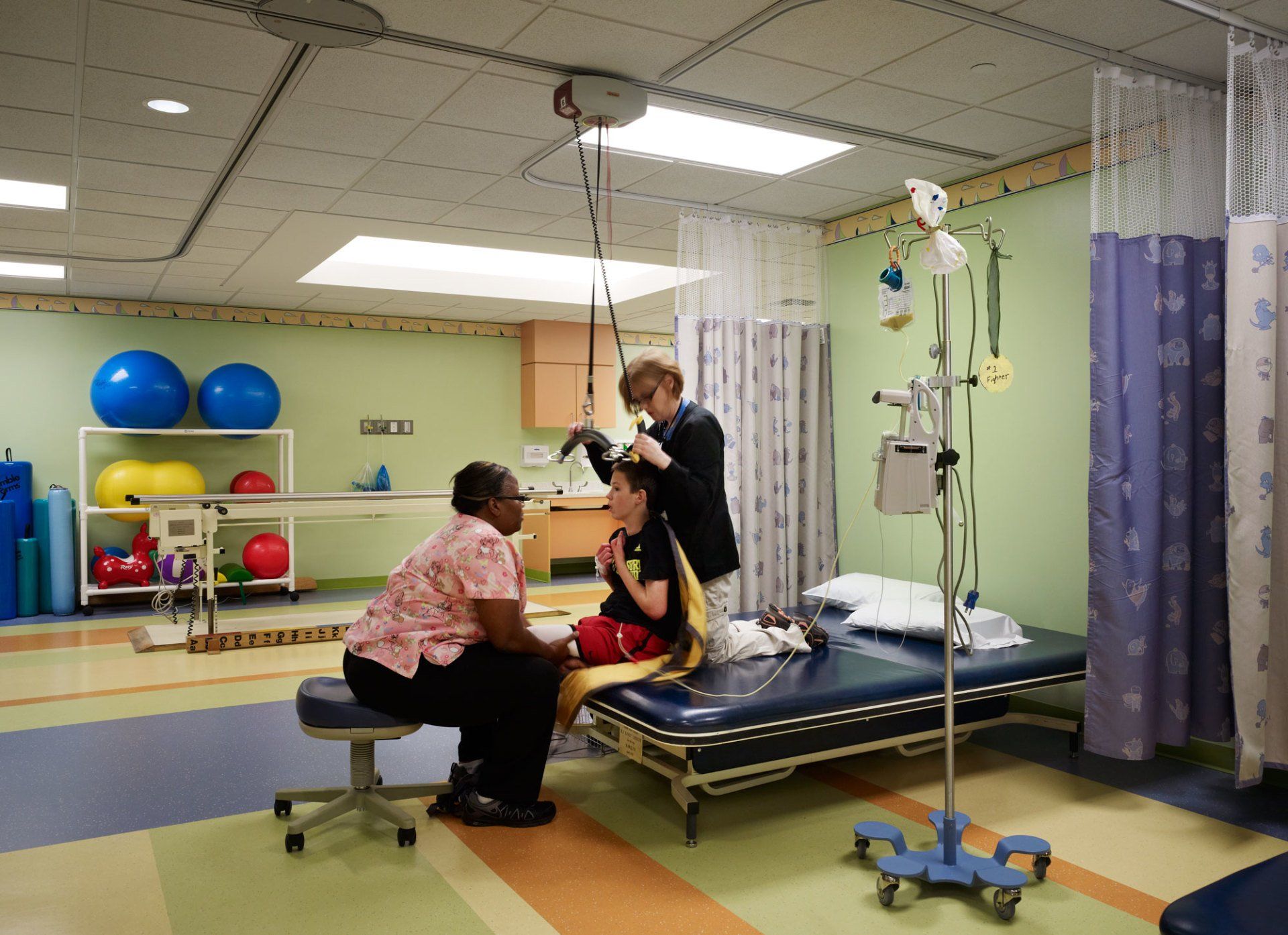 Two healthcare workers attend to a child lying on a hospital bed; therapy equipment is in the background.