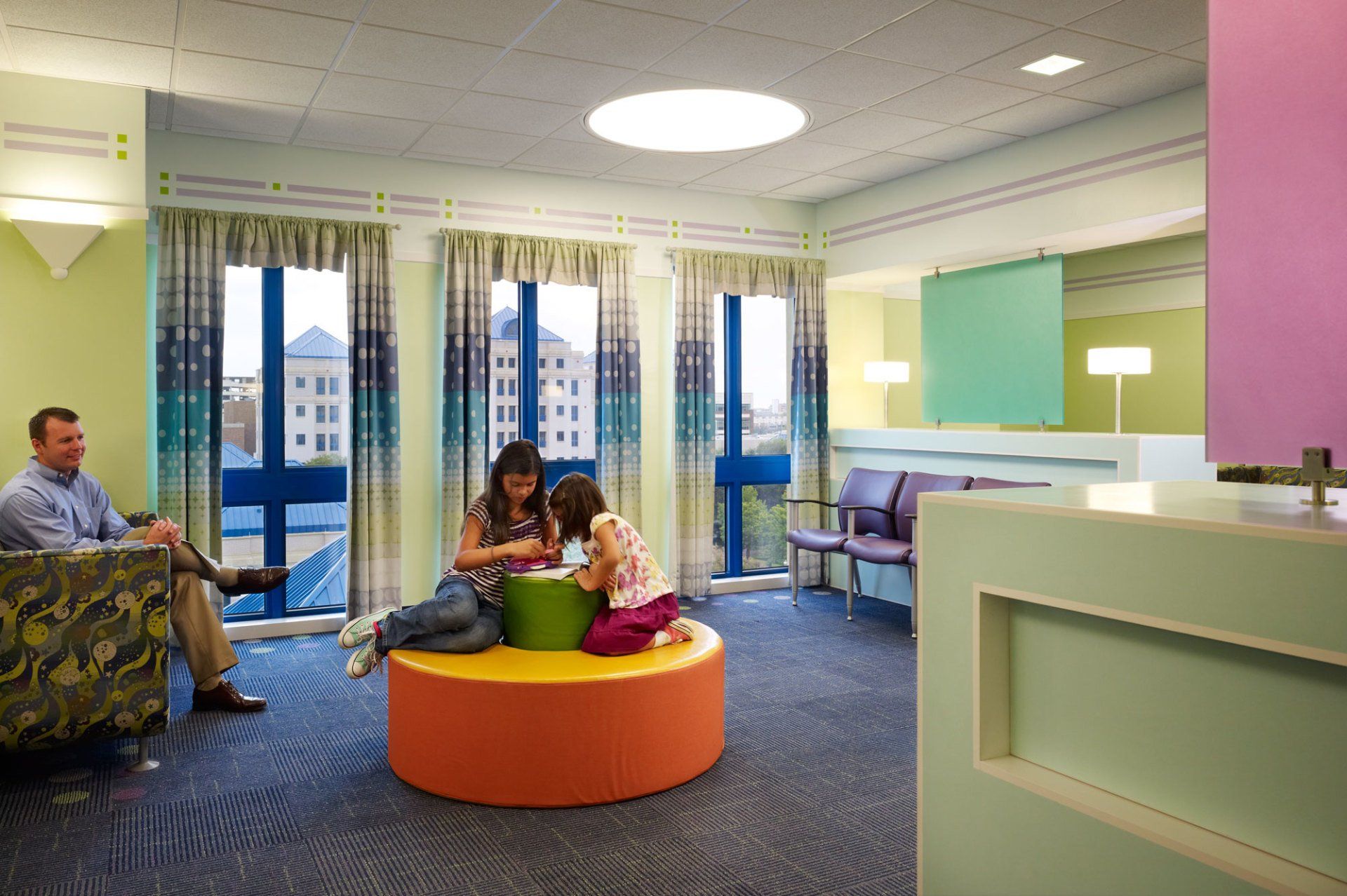 A colorful waiting room with a family: a man seated, a mother and child playing on a round orange and green seat. Blue windows and walls, various colorful accents.
