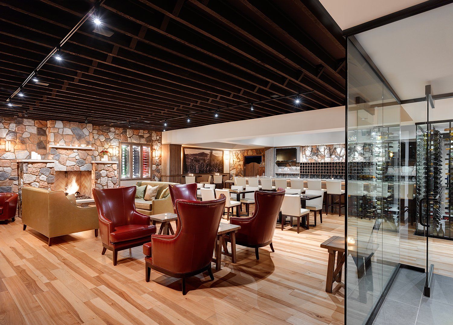 Interior of a stylish restaurant with a stone wall, leather chairs, a bar, and a glass-enclosed wine cellar. The lighting is warm and inviting.