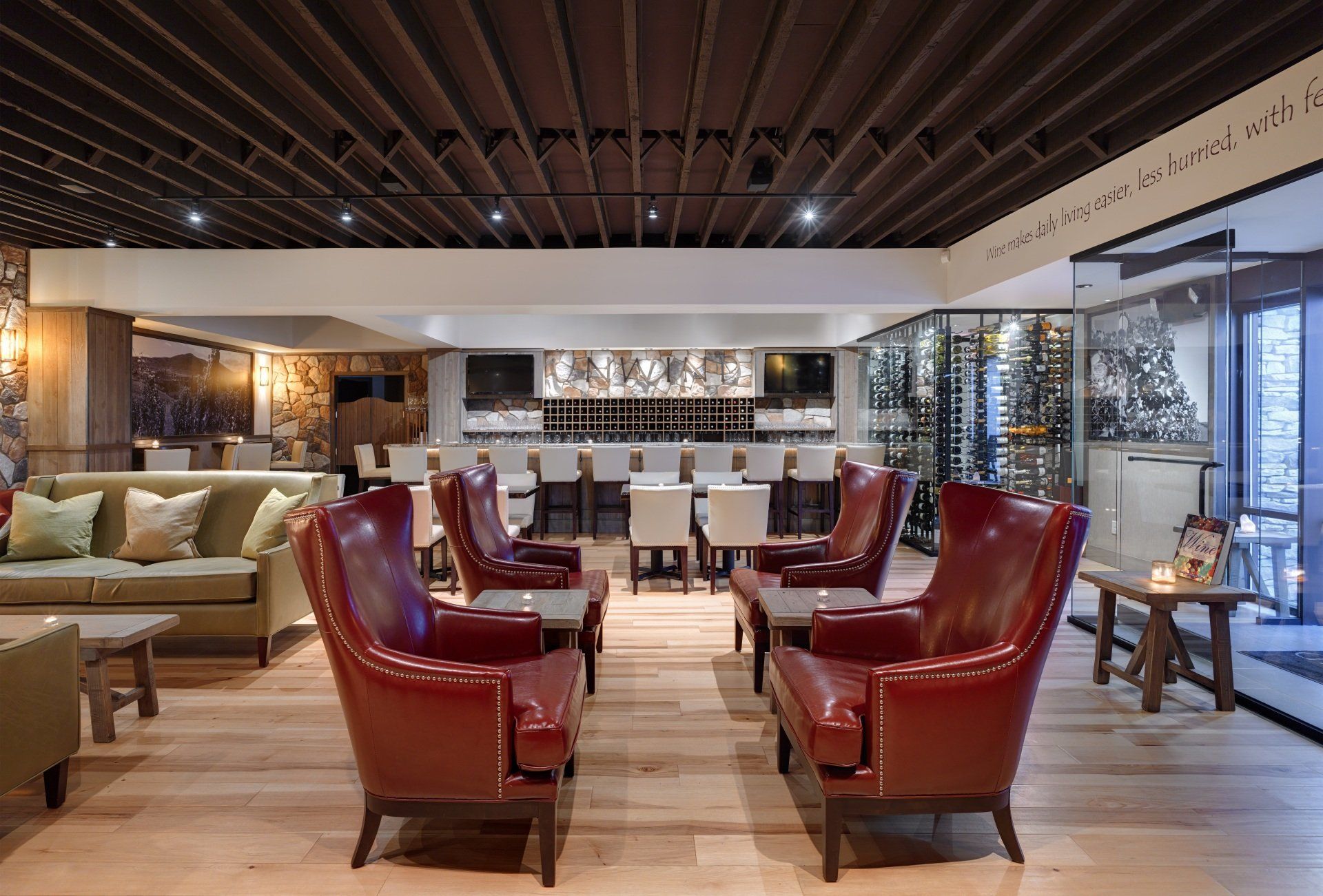 A lounge with red leather armchairs, a bar with white stools, and a wine display. The setting has wooden floors and a stone wall.