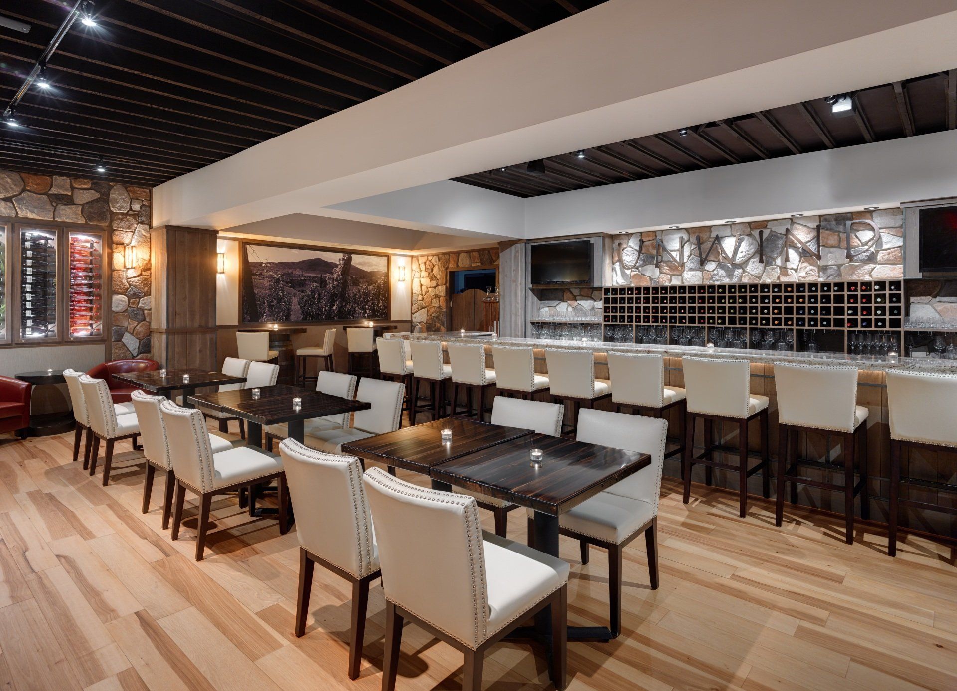 Restaurant interior featuring a bar with stools and tables with chairs. Walls are stone and wood. The floor is light wood.