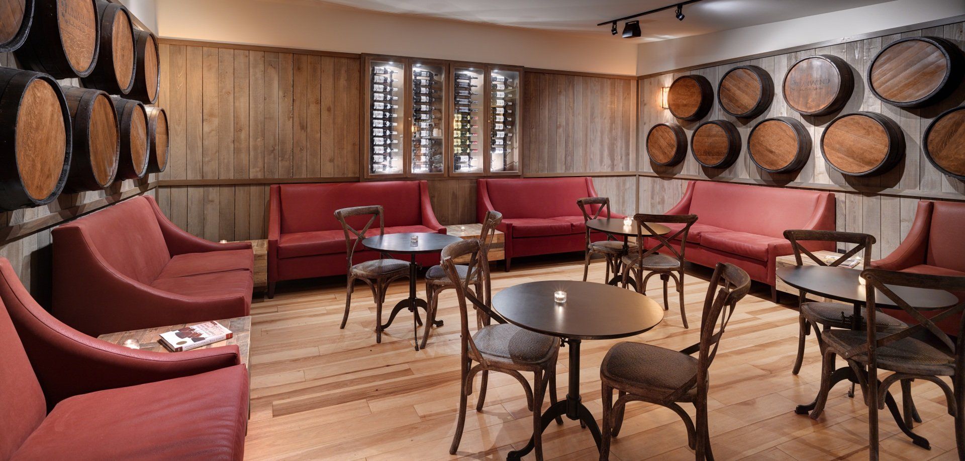 Cozy lounge with wooden barrels, red sofas, small round tables, and chairs, creating a wine-tasting atmosphere.