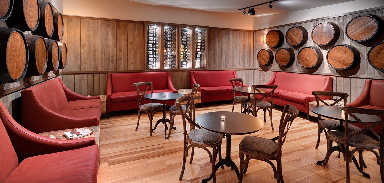 Cozy wine cellar with red couches, wooden tables, and chairs, and barrels lining the walls.
