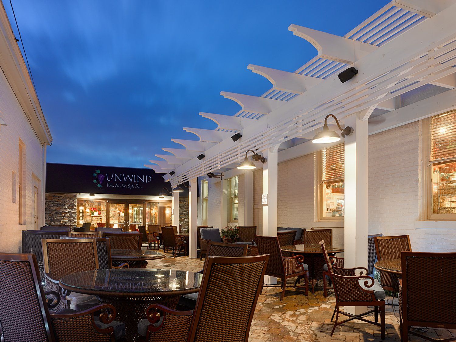 Outdoor patio with wicker furniture, beneath a white pergola lit by overhead lamps. Restaurant entrance visible in the background.