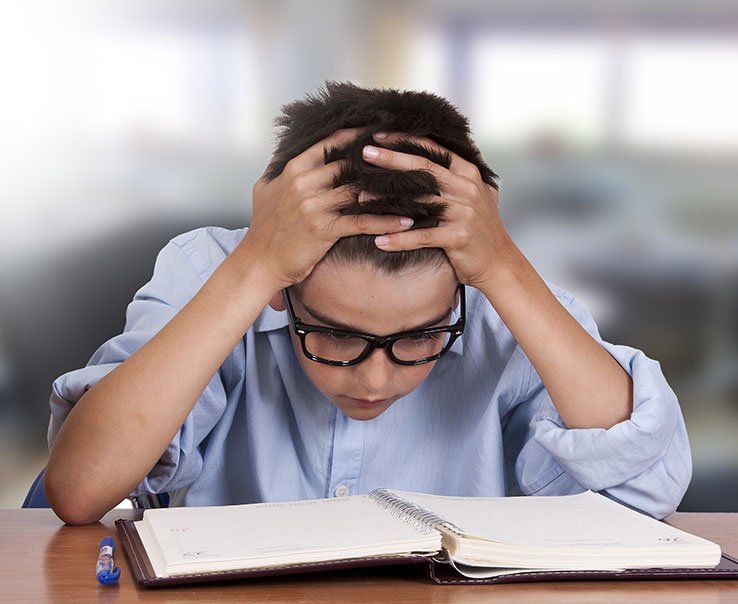 Un garçon portant des lunettes est assis à un bureau avec ses mains sur la tête.