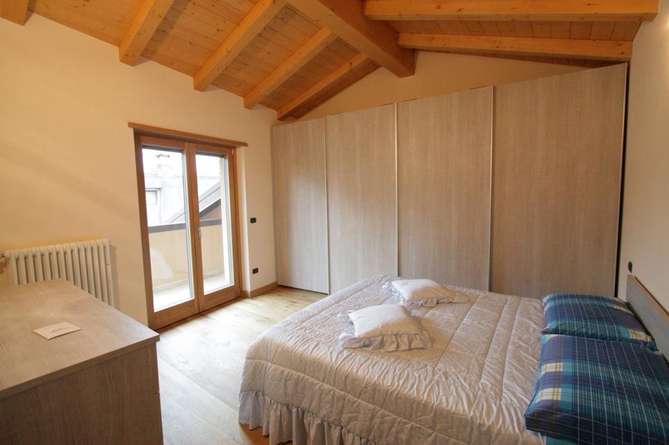 Accogliente camera da letto mansardata con letto, armadio, cassettiera e soffitto in legno, accessibile tramite porte finestre.
