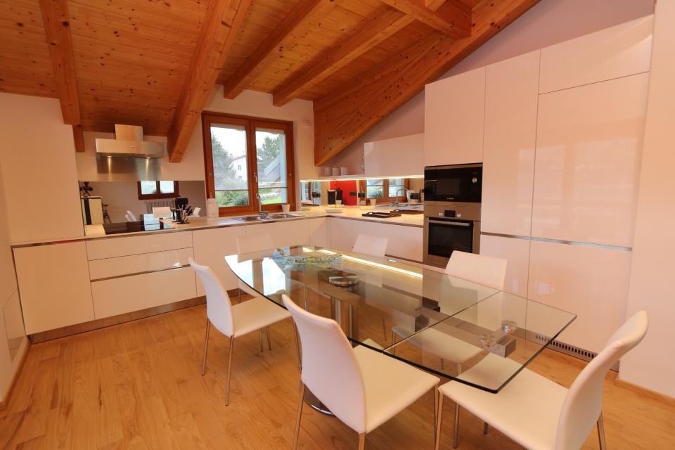 Cucina e zona pranzo moderne con soffitto in legno, tavolo in vetro e sedie bianche.