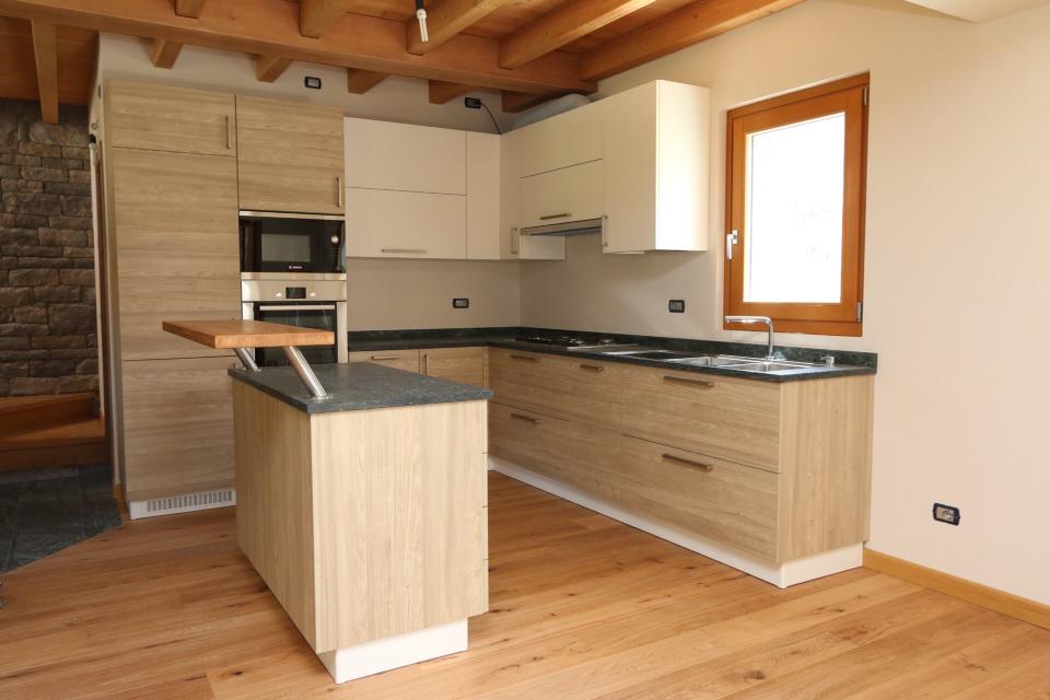 Cucina moderna con mobili in legno chiaro, ripiani neri, isola e un'ampia finestra sotto un soffitto con travi in legno.