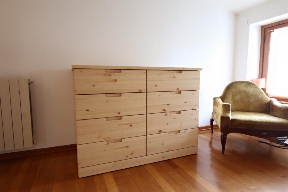 Cassettiera in legno in una stanza soleggiata accanto a una poltrona verde e alla finestra.
