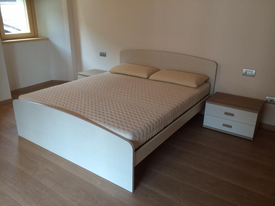 Camera da letto moderna con letto beige, due cuscini e comodino abbinato su pavimento in legno.