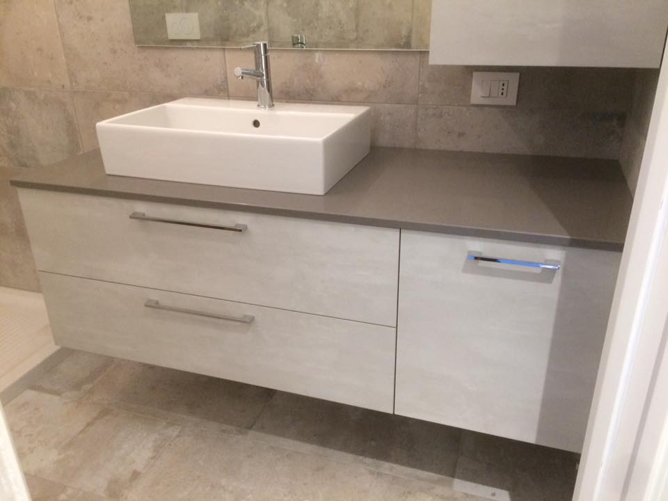 Mobile bagno moderno con lavabo bianco, piano grigio e ante in legno chiaro a contrasto con le pareti piastrellate.