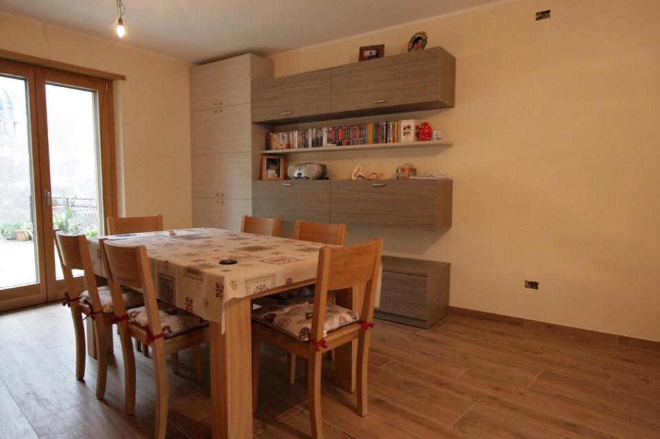 Sala da pranzo con tavolo e sedie in legno, mensole a muro e una finestra che lascia entrare la luce naturale.