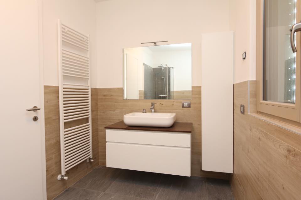 Bagno moderno con mobile lavabo bianco, specchio, termosifone e pareti rivestite con piastrelle beige.