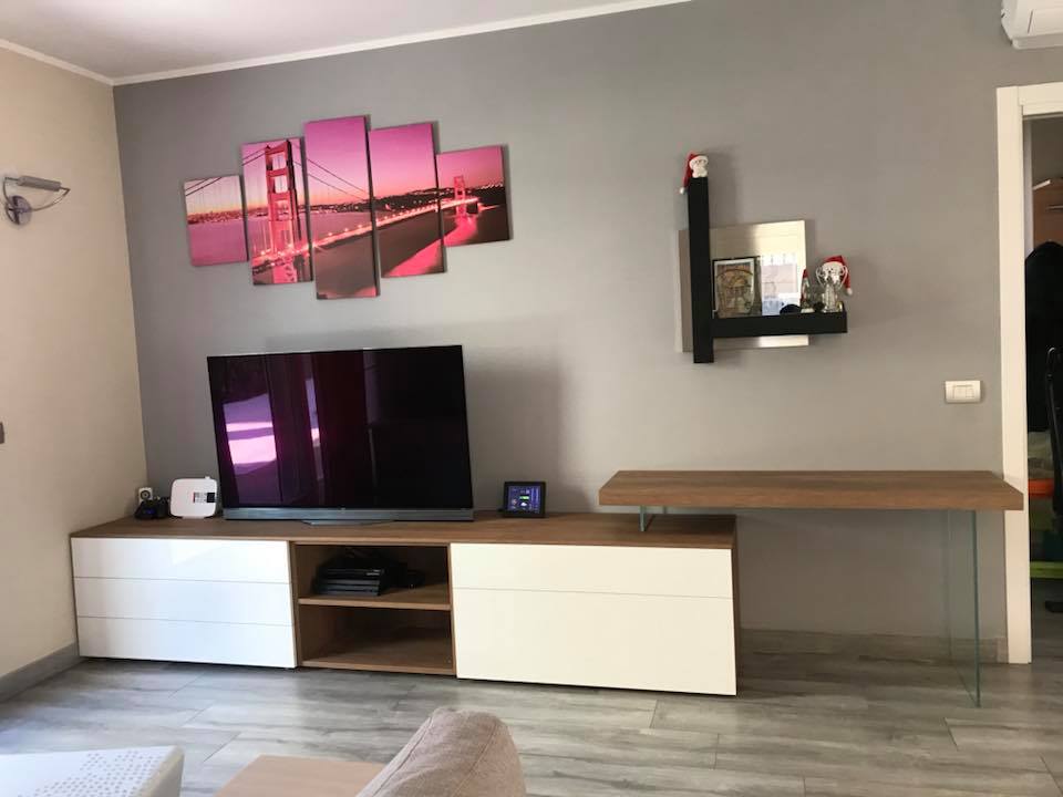 Soggiorno moderno con TV su mobile porta TV bianco, quadri rosa alle pareti, pareti grigie e mensole a vista.