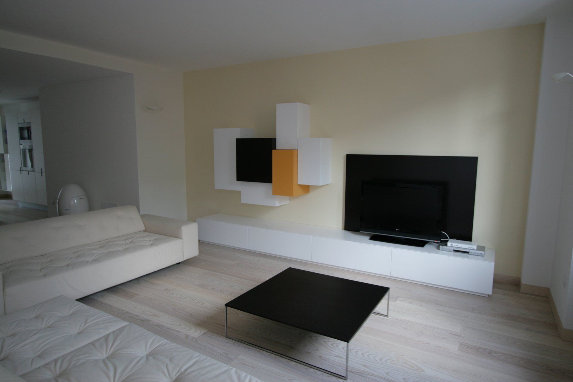 Soggiorno moderno e minimalista con divano componibile bianco, TV a parete e tavolino nero su pavimento chiaro.