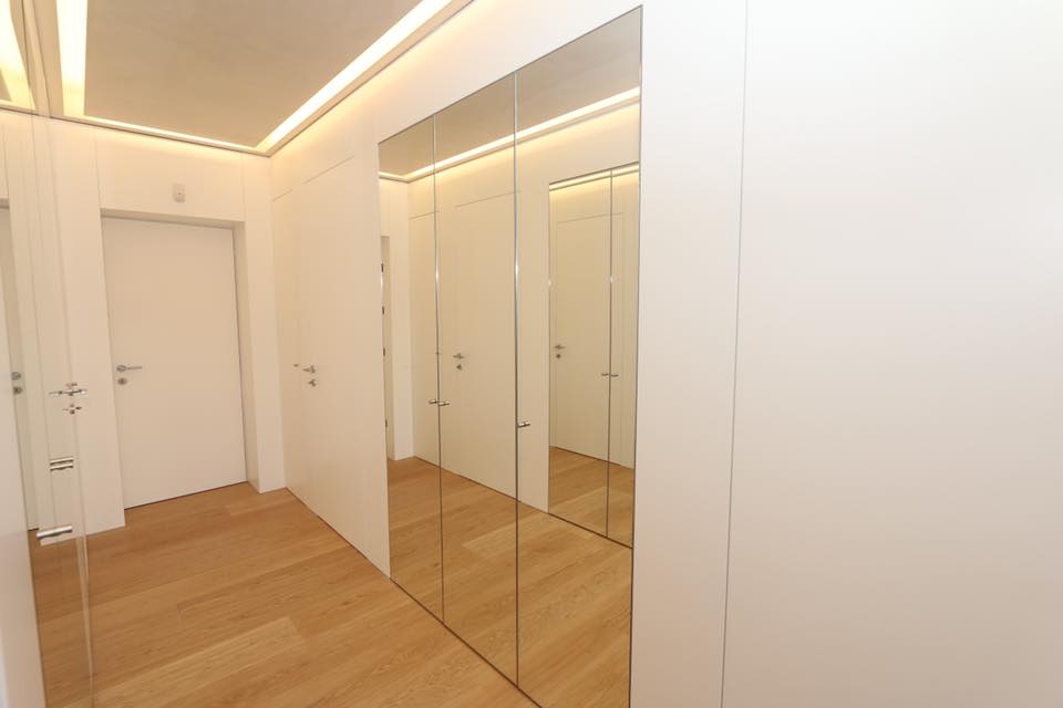 Corridoio moderno con pavimento in legno, ante dell'armadio a specchio, pareti bianche e faretti a incasso sul soffitto.