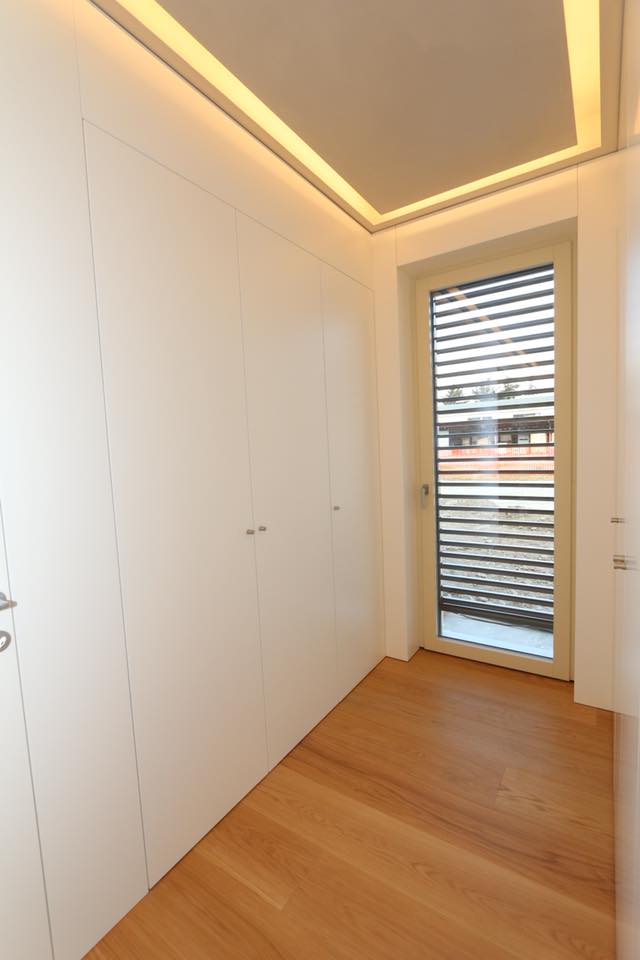 Luminoso corridoio minimalista con armadi a muro bianchi, pavimento in legno e porta finestra con tende.