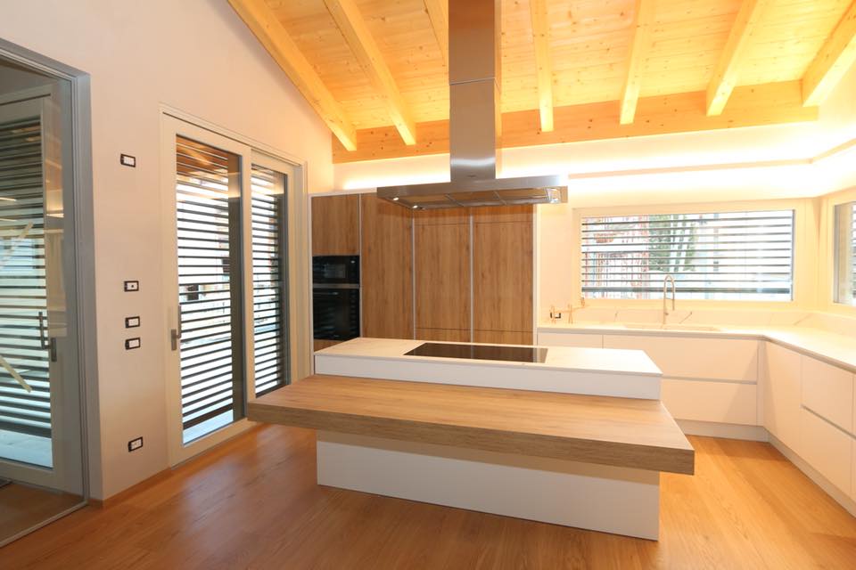 Cucina moderna con mobili in legno, ripiani bianchi e isola centrale illuminata da una luce calda.