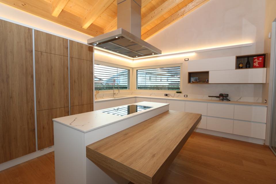 Cucina moderna con dettagli in legno, mobili bianchi, isola e cappa aspirante in acciaio inox sotto un soffitto inclinato in legno.