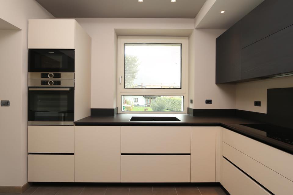 Cucina moderna e minimalista con mobili bianchi, ripiani neri, forno a incasso e finestra sopra il lavello.