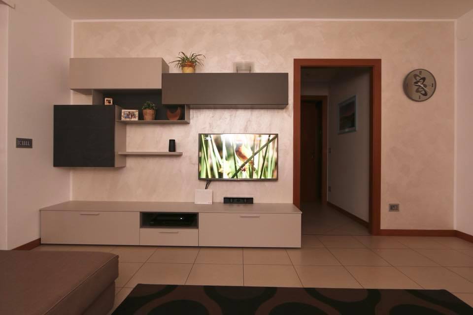Soggiorno moderno con TV a parete, mobile porta TV bianco, armadi scuri e pavimento piastrellato beige.
